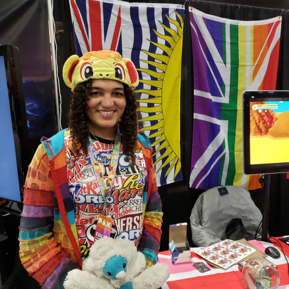 Rina Purdy stands near a colorful booth with a brightly-colored hat and rainbow-colored shirt.